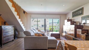 Un vaste espace de vie comprenant un canapé, un pouf et une table à manger avec des chaises. Un escalier mène à l’étage supérieur, et on y trouve des placards et un radiateur sur le côté droit.
