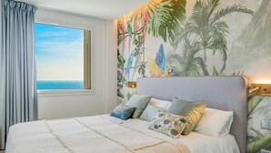 A bedroom with a large bed, a view of the sea through a window, and a tropical mural on the wall. - Tropical Suite by Hello Homes Sitges (Sitges)