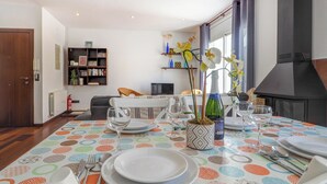 Un comedor con una mesa dispuesta para cuatro personas, rodeada de sillas. Se observa una estantería y una chimenea al fondo.