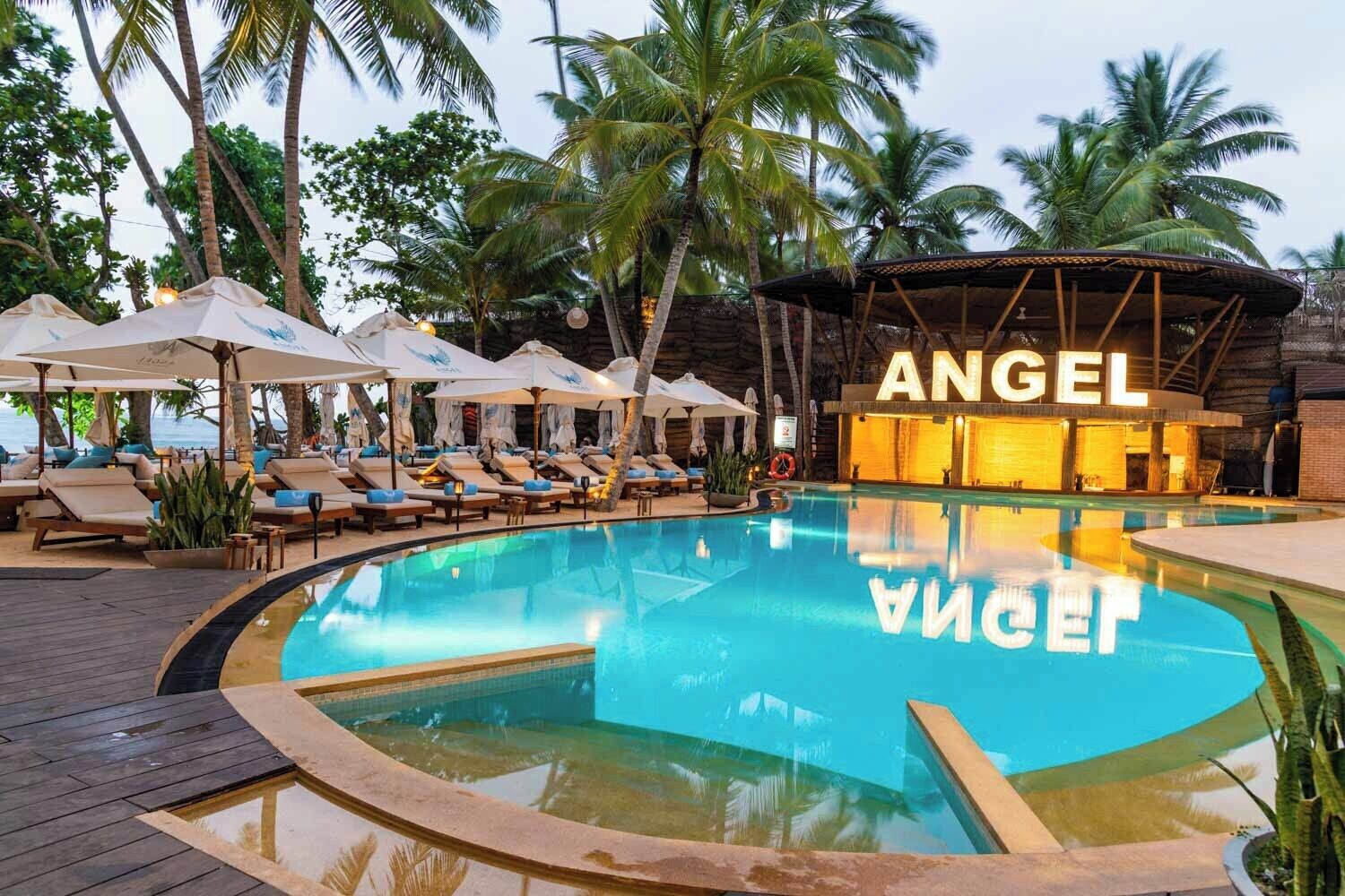 Un área de piscina con sillas de descanso y sombrillas, un edificio con la etiqueta 'ANGEL', y palmeras.