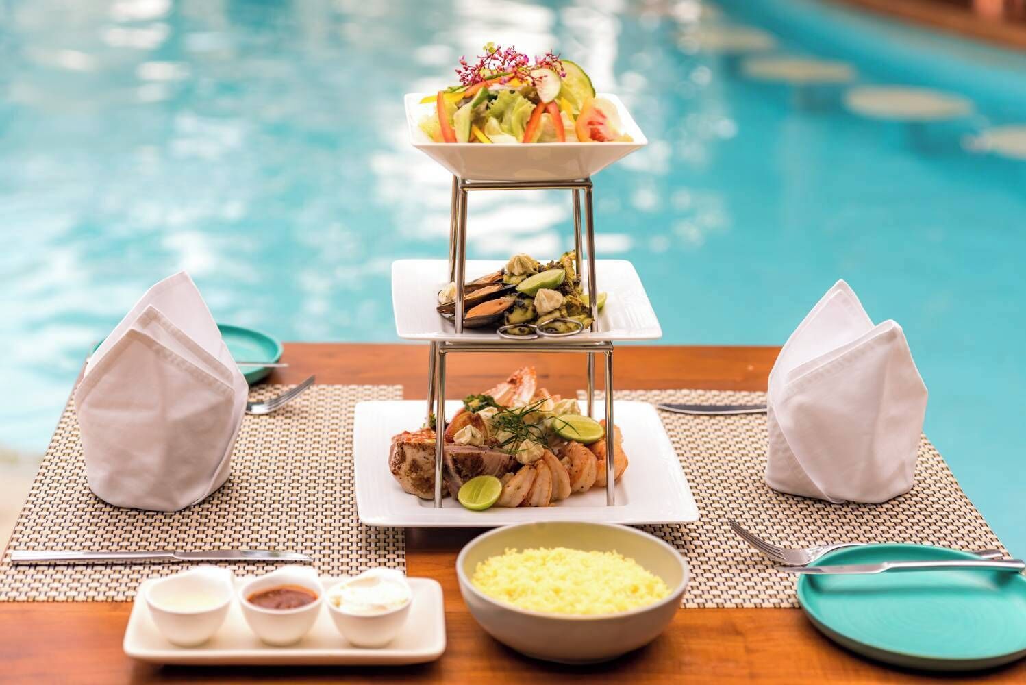 Montaje de comedor con vista a la piscina, que incluye un expositor de comida de tres niveles, servilletas, cubiertos y condimentos.