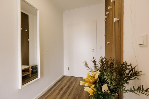 A hallway with a wooden floor, a white door, and a mirror reflecting a room with a chair and a dog bed. - De Diek - Suite 4 | Lateral dike location (Greetsiel)