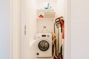 Une petite buanderie comprenant un lave-linge, une étagère avec des serviettes et une trousse de premiers secours, ainsi qu’un balai appuyé contre le mur.