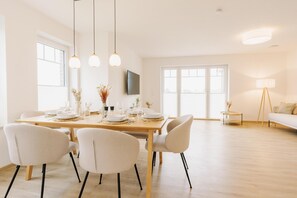A modern dining area with a wooden table set for four, white chairs, and pendant lighting. - Canals - Suite 1 | Direct Waterfront Location (Greetsiel)