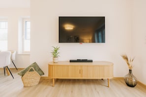 A modern living room with a flat-screen TV mounted on the wall, a wooden console table, a wicker basket, a potted plant, and a vase with dried plants.