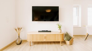 A modern living room with a flat-screen TV mounted on the wall, a wooden console table, a vase with dried plants, and a wicker basket. - Canals - Suite 2 | Direct Waterfront Location (Greetsiel)