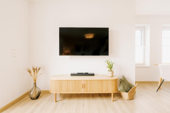 A modern living room with a flat-screen TV mounted on the wall, a wooden console table, a vase with dried plants, and a wicker basket. - Canals - Suite 2 | Direct Waterfront Location (Greetsiel)
