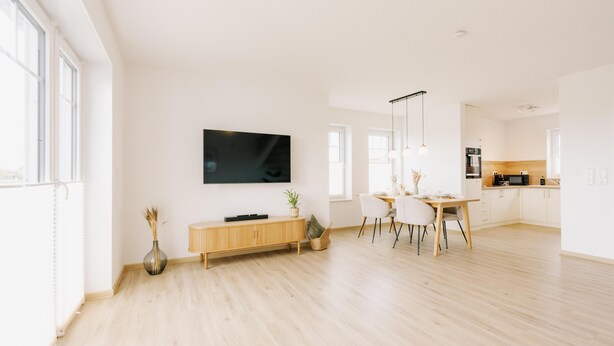 A modern living room with a flat-screen TV, wooden flooring, a dining area with chairs, and a kitchen in the background. - Canals - Suite 2 | Direct Waterfront Location (Greetsiel)