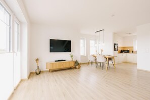 A modern living room with a flat-screen TV, wooden flooring, a dining area with chairs, and a kitchen in the background. - Canals - Suite 2 | Direct Waterfront Location (Greetsiel)