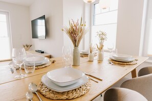 A dining table set with plates, glasses, and cutlery, with a vase of dried plants and a wall-mounted TV. - Canals - Suite 2 | Direct Waterfront Location (Greetsiel)