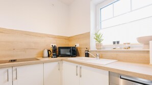A modern kitchen with white cabinets, a stainless steel dishwasher, a microwave, a kettle, and a window with a plant. - Canals - Suite 2 | Direct Waterfront Location (Greetsiel)