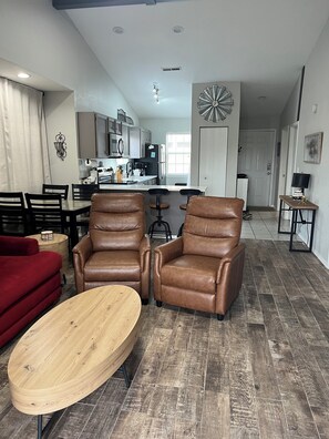 Ein modernes Wohnzimmer mit Ledersesseln, einem Holztisch und einem roten Sofa. Im Hintergrund ist eine Küche mit Edelstahlgeräten zu sehen.