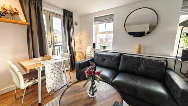 A modern living room with a black leather sofa, a round coffee table, a dining table with chairs, a small side table with a vase of flowers, and a large mirror on the wall.