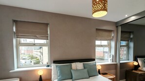 A modern bedroom with a large bed, two bedside tables, a nightstand, and a window with a view of buildings.
