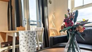 A cozy room with a round wooden table, a chair with a patterned cloth, a sofa, and a vase with flowers on a windowsill.