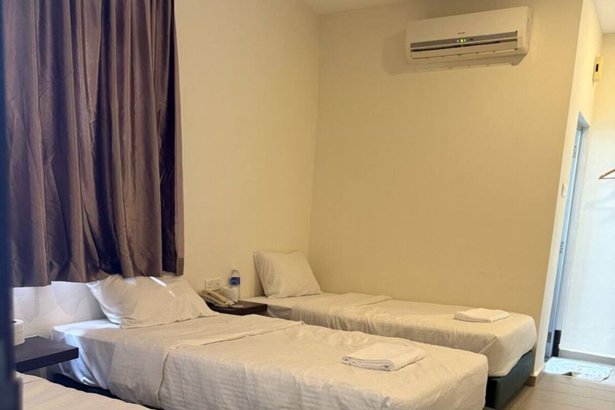 A hotel room with three beds arranged in a row, a nightstand with a lamp, an air conditioning unit on the wall, and a window with curtains.