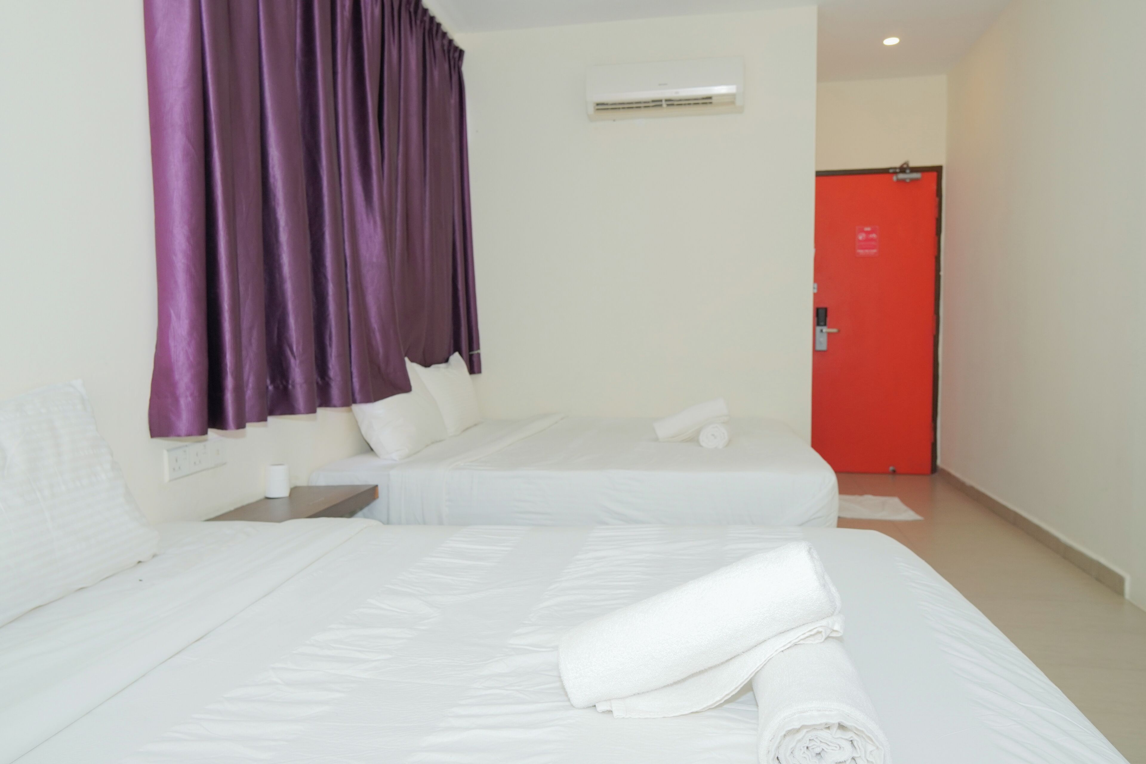 A hotel room with two beds, purple curtains, a red door, and an air conditioning unit.