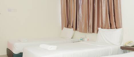 A hotel room with three beds, each with white linens and pillows. There is a bedside table with a lamp and a phone. The room has a large window with brown curtains.