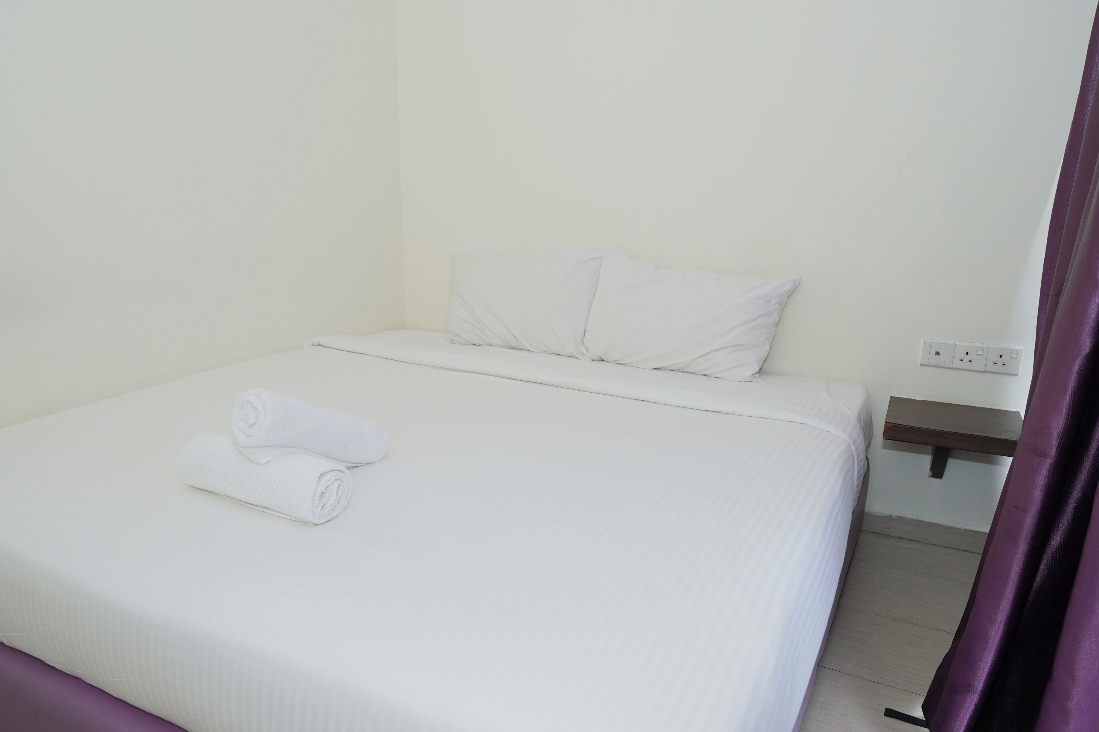 Una cama bien hecha con sábanas blancas y una toalla enrollada, una mesita de noche de madera con un enchufe, y una cortina morada parcialmente visible.