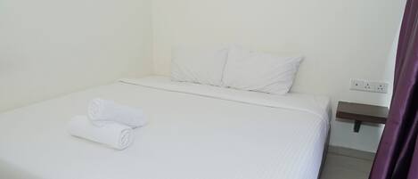 Una cama bien hecha con sábanas blancas y una toalla enrollada, una mesita de noche de madera con un enchufe, y una cortina morada parcialmente visible.