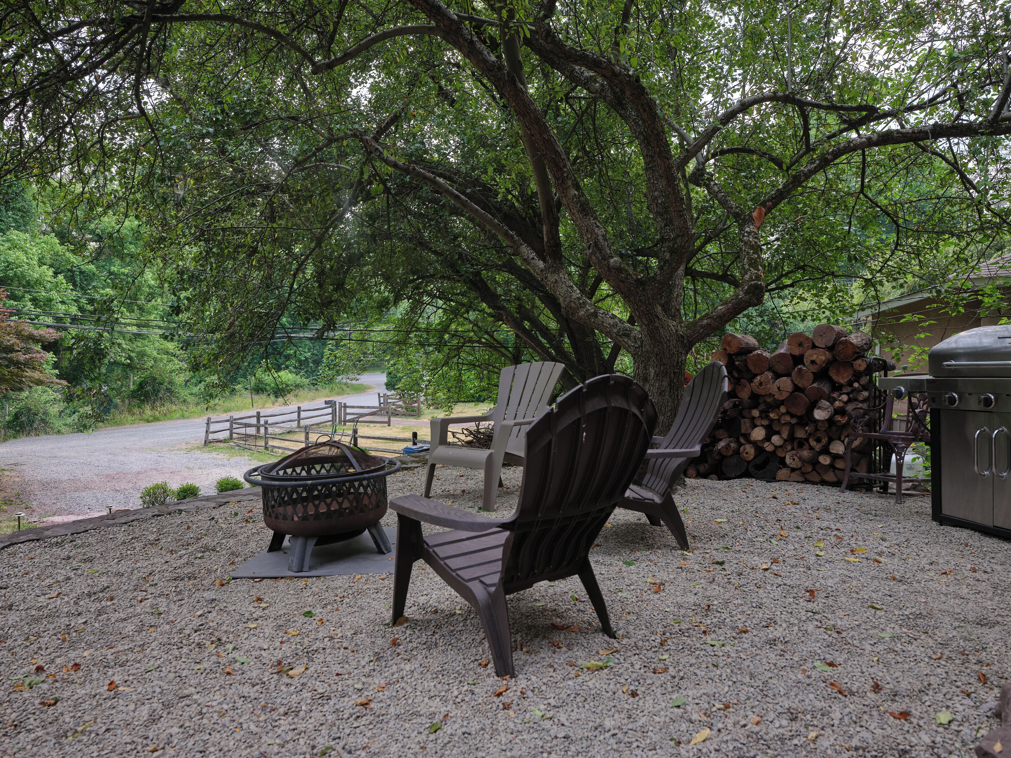 A gravel patio with Adirondack chairs, a fire pit, and a grill area surrounded by trees.