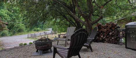 A gravel patio with Adirondack chairs, a fire pit, and a grill area surrounded by trees.