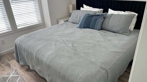 Un lit bien fait, avec une couverture grise à texture et des oreillers, une tête de lit sombre et une petite table de chevet avec une lampe.