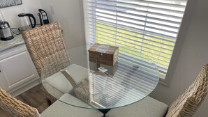 Une table ronde en verre, des chaises en osier, une boîte étiquetée « EMBALLÉ AVEC LES DONS DE LA NATURE » et une fenêtre avec des stores.
