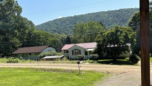 Entorno rural con una casa de techo rojo y un pequeño porche, rodeada de exuberante vegetación y una colina al fondo.