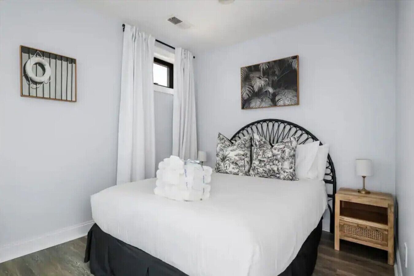 Une chambre à coucher avec un lit, des rideaux blancs, une fenêtre, un tableau encadré et une table de chevet avec une lampe.