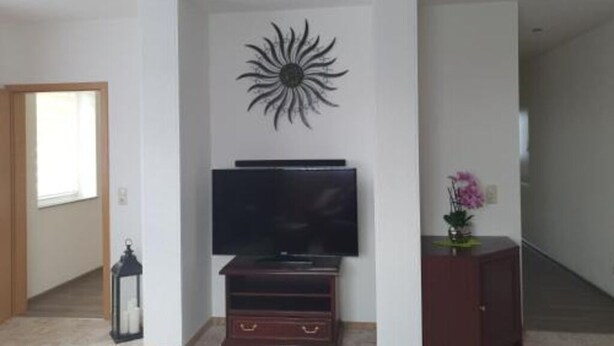 Un salon avec un téléviseur à écran plat posé sur un meuble en bois, une décoration murale représentant un soleil, une petite table avec un vase, et un escalier en arrière-plan.