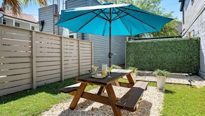 Una mesa de picnic de madera con un paraguas azul, rodeada por un área de grava y una valla de madera.