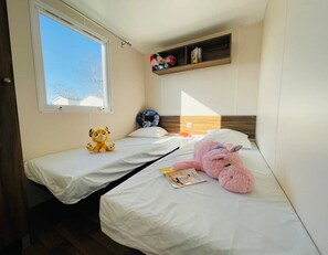 A small, well-lit room with two beds, a window, and a shelf with plush toys.