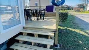 Une terrasse en bois mène à un espace extérieur abrité, équipé d’une table et de chaises, d’un cerceau en plastique bleu et d’une zone herbeuse avec un sentier.
