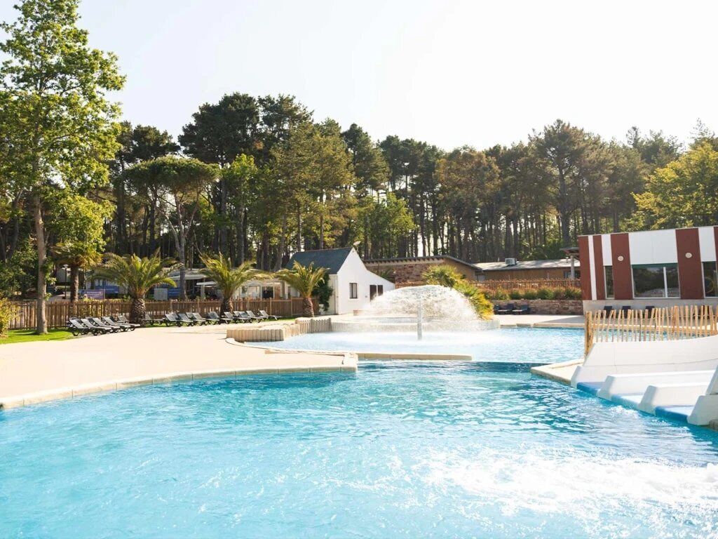 A swimming pool with a water fountain, surrounded by lounge chairs and a small building.