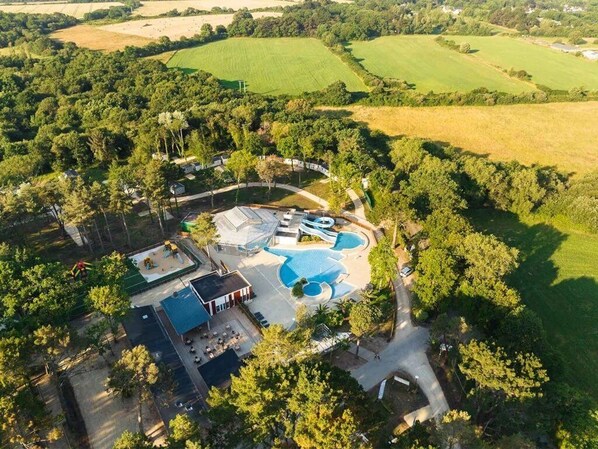 An aerial view of a resort with a swimming pool, tennis courts, and a parking area surrounded by trees and fields.