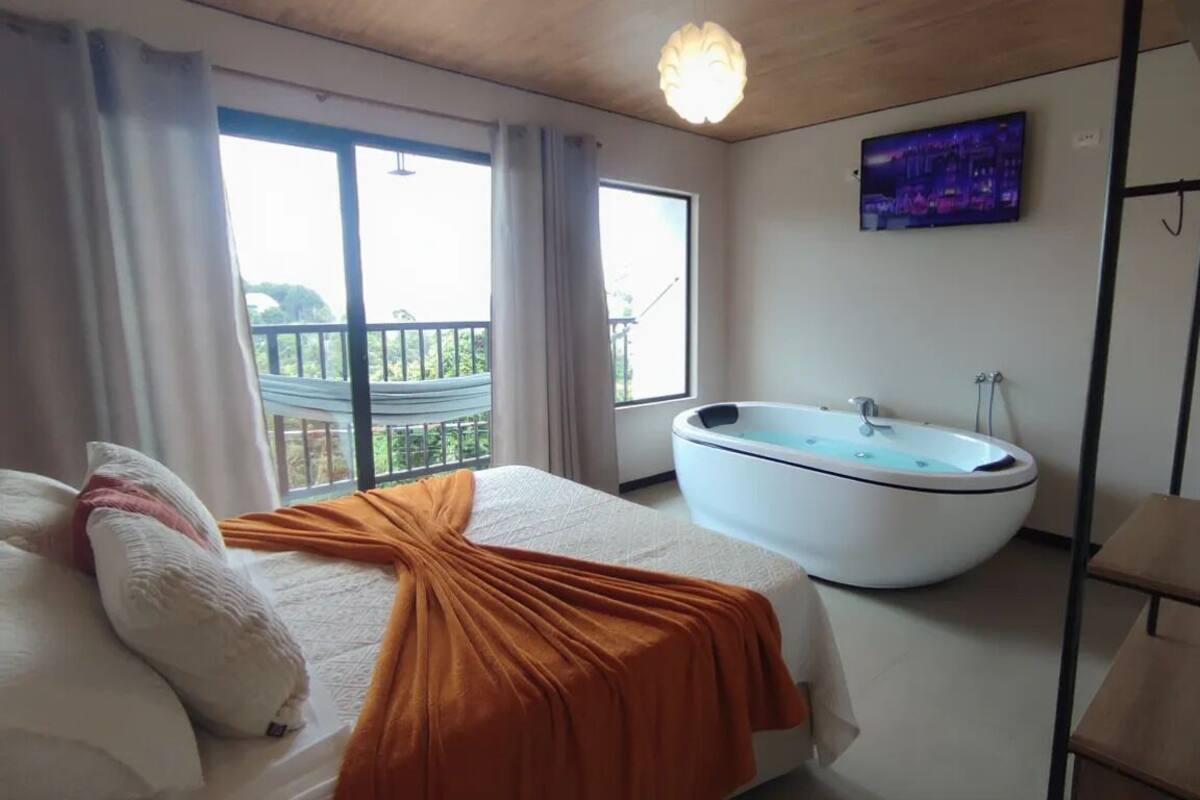 Ein Schlafzimmer mit einem großen Bett, einem eigenen Badezimmer mit Badewanne, einem Balkon mit Geländer und einem an der Wand befestigten Fernseher.