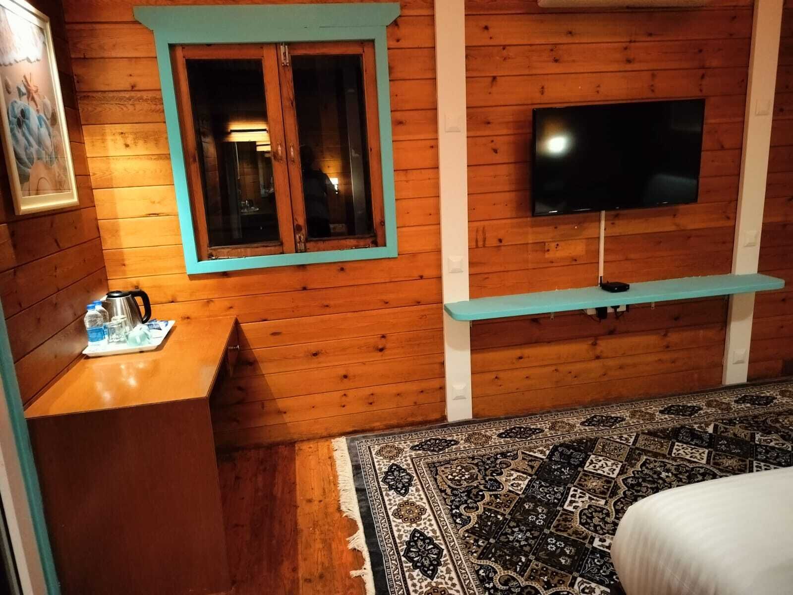 Una habitación de cabaña de madera con un televisor montado en la pared, una mesita con una tetera y una alfombra estampada en el suelo.