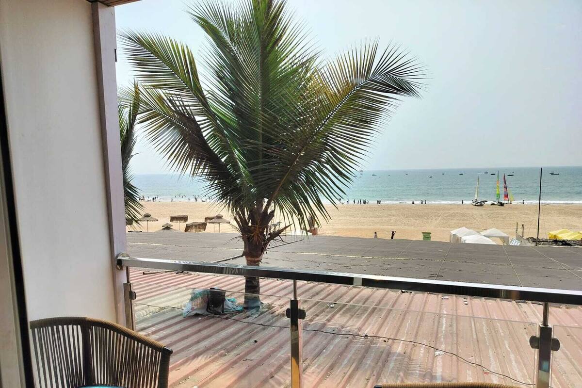 A balcony with a view of a beach, a palm tree, and two wicker chairs.