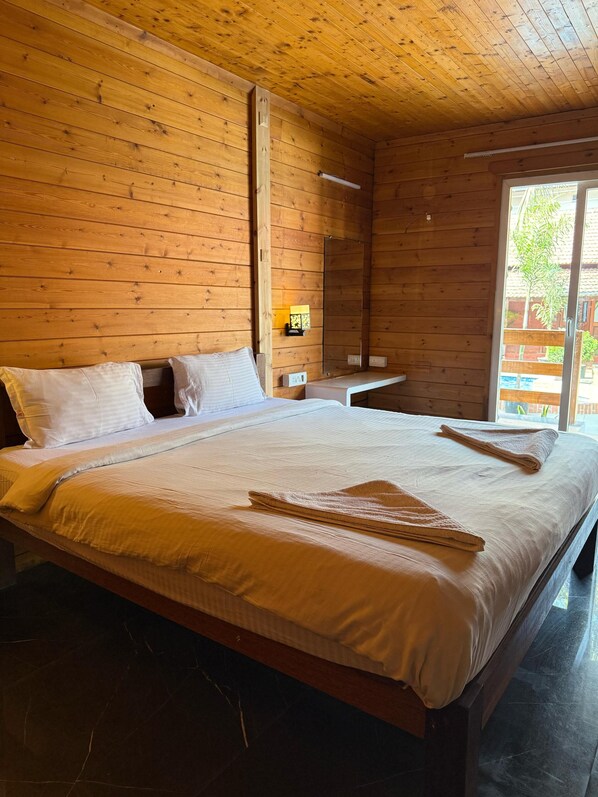 Une chambre à coucher aux murs lambrissés, avec un grand lit, une table de chevet, une lampe et une porte-fenêtre coulissante donnant sur l’extérieur.