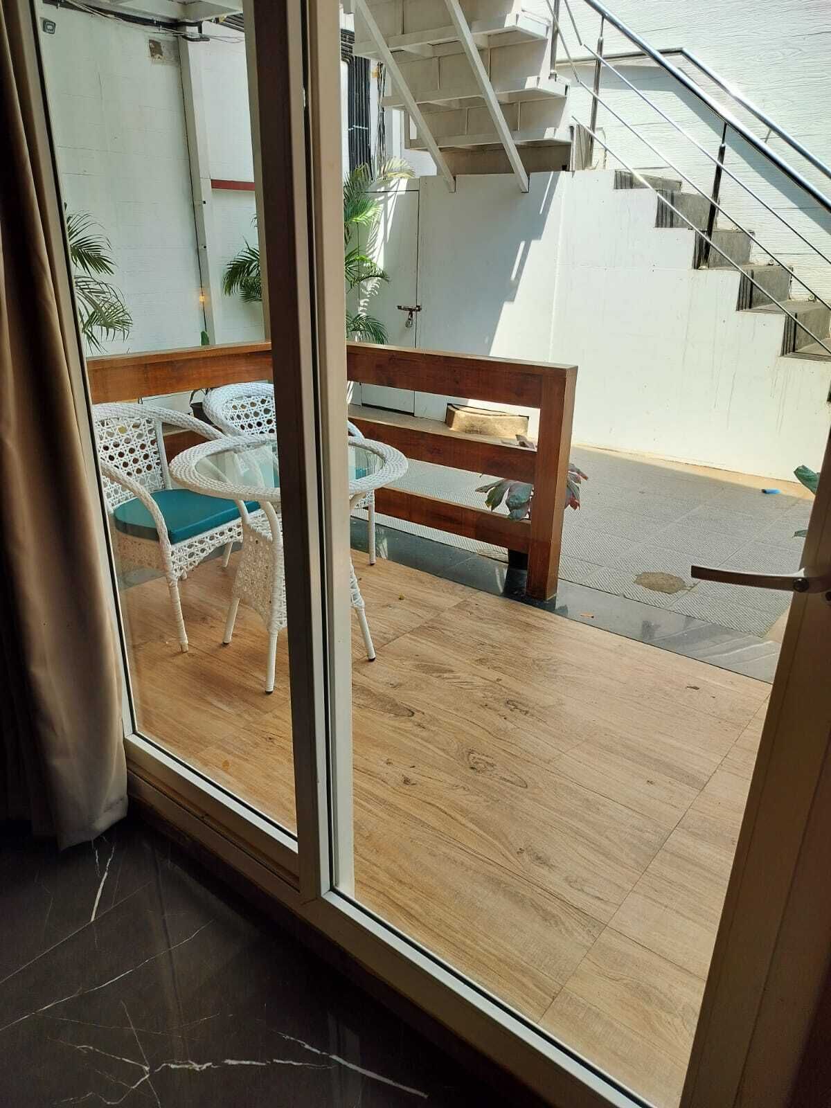 A room with a sliding glass door leading to an outdoor area with a wooden deck, a white wicker table with blue cushions, and a wooden bench.