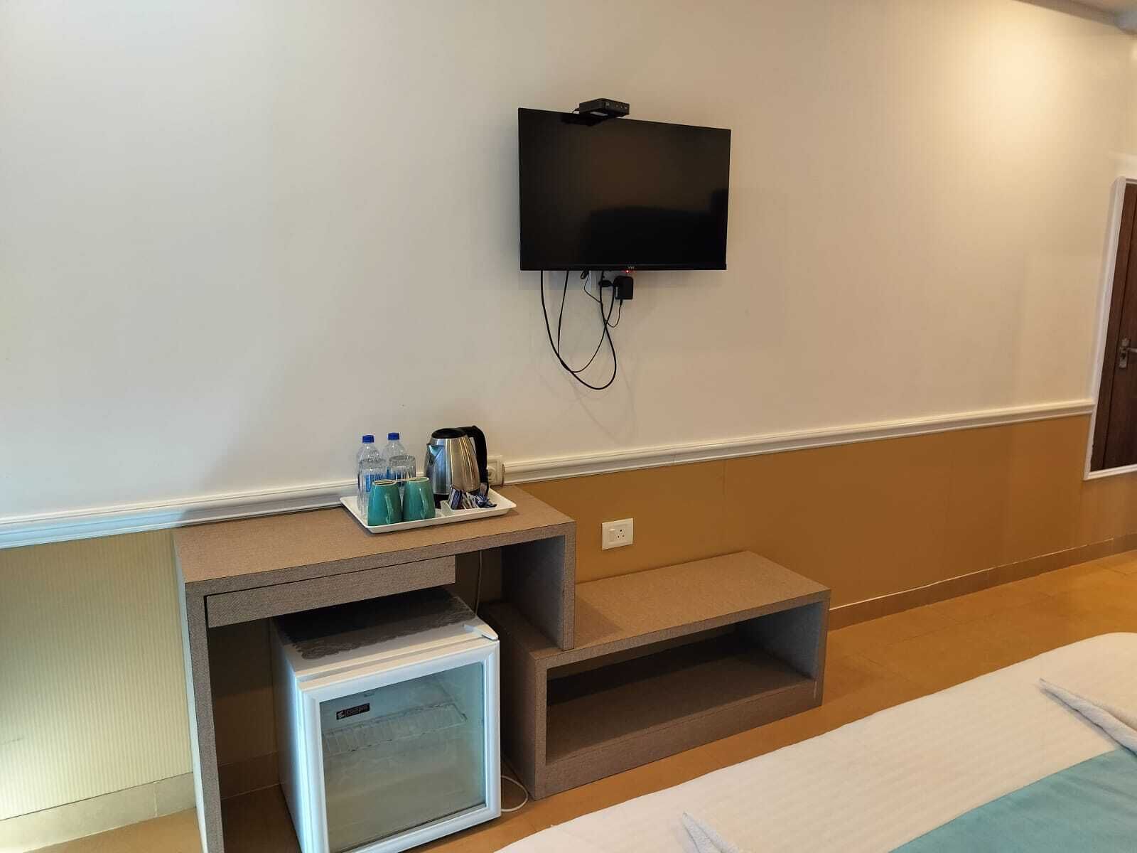 A hotel room with a bed, a small refrigerator, a TV mounted on the wall, and a tray with bottles and cups.