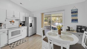 Una cocina moderna con armarios blancos, una estufa y un refrigerador. Hay un comedor con una mesa y sillas blancas, y vistas al exterior a través de una puerta corrediza de vidrio.