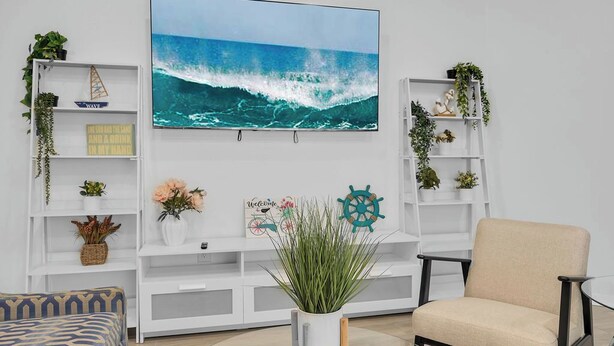 Sala de estar con una televisión grande montada en la pared que muestra una ola, un sillón color beige, un sofá estampado y estanterías blancas con plantas y objetos decorativos.