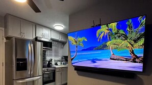 A modern kitchen with stainless steel appliances, a flat-screen TV mounted on the wall, and a small wooden table with a decorative plate. - Welcome to SD Cozy Home - B (La Mesa)