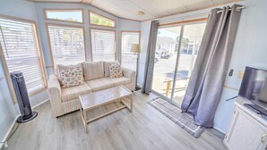 Una sala de estar acogedora con un sofá, una mesa de centro y un televisor. Amplias ventanas con persianas, un radiador y una puerta que da al exterior.