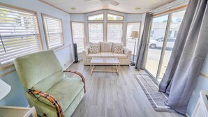Una habitación con un sillón verde, un sofá a rayas, un ventilador de pie y ventanas grandes con persianas.