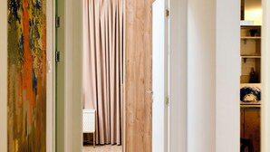 A hallway with wooden flooring, a door leading to a room with a bed and curtains, and a chair in the corner.