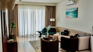 A modern living room with a sofa, two armchairs, a coffee table, and a desk with a telephone. A painting of a seascape hangs on the wall, and an air conditioning unit is mounted above the sofa.
