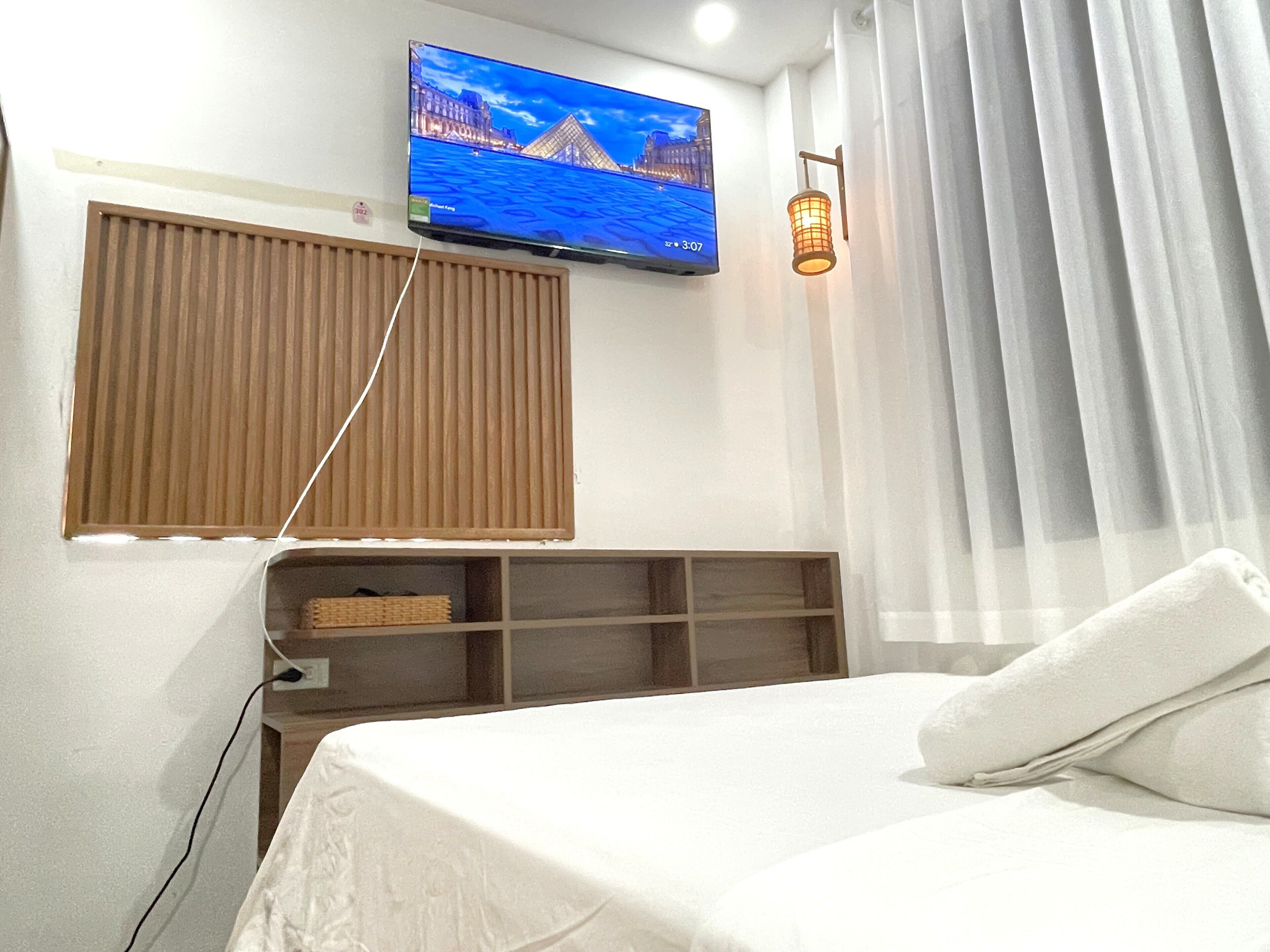 A hotel room with a bed, a TV mounted on the wall, a window with blinds, a shelf with a basket, and a wall-mounted lamp.